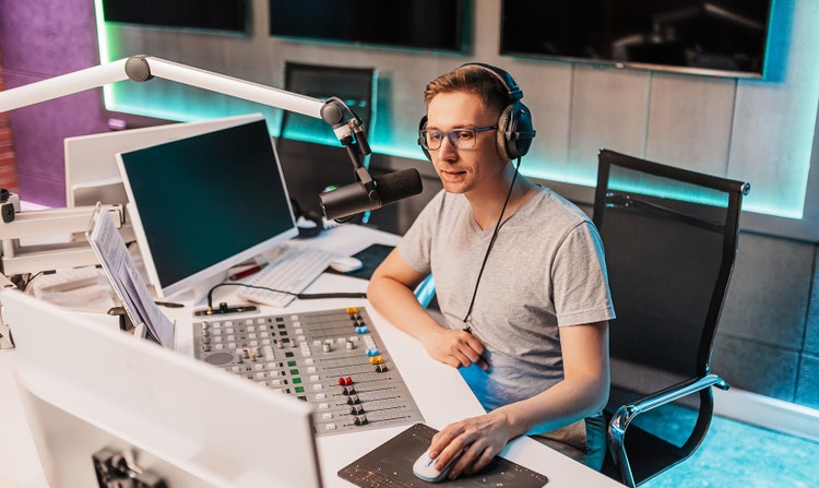 A person recording at a professional audio workstation with a microphone, mixing console, and computer monitors. The workspace illustrates creating high-quality AI voiceovers efficiently for podcasts, videos, and other content at scale.