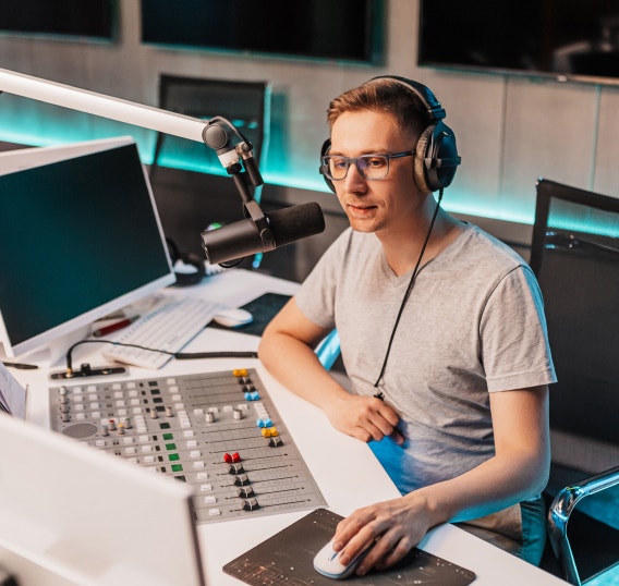 A person recording at a professional audio workstation with a microphone, mixing console, and computer monitors. The workspace illustrates creating high-quality AI voiceovers efficiently for podcasts, videos, and other content at scale.