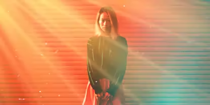 A person staring at the camera with hands held in front of them is bathed in orange and yellow rays of light