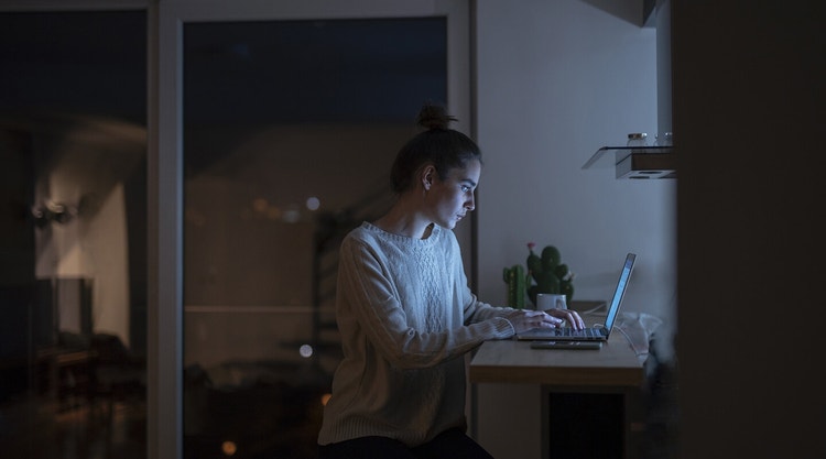 Uma pessoa a elaborar uma carta de motivação no portátil, sentada num balcão de cozinha no escuro