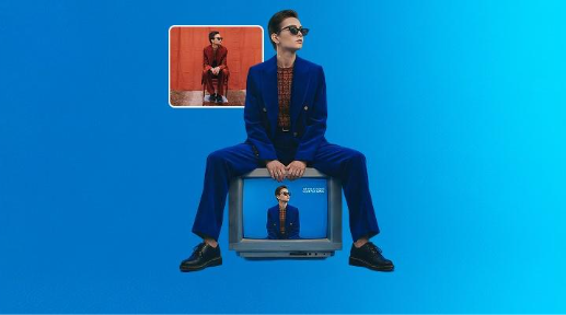 A woman in an orange suit sits on a chair in a field. The Photoshop AI Assistant is used to swap in a blue background, to replace the chair with TV and change the suit's colour to blue.