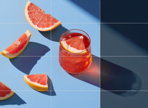 O fotografie cu felii de grepfrut lângă un cocktail roz este extinsă pentru a arăta umbra completă proiectată de pahar.