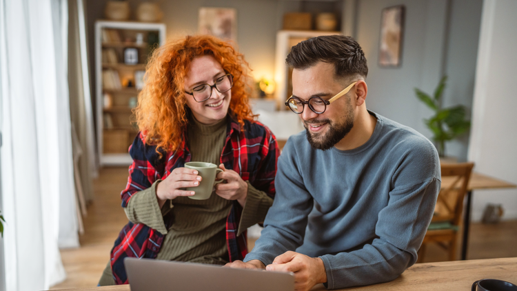 A redhaired woman and a dark haired man smile and look at a laptop