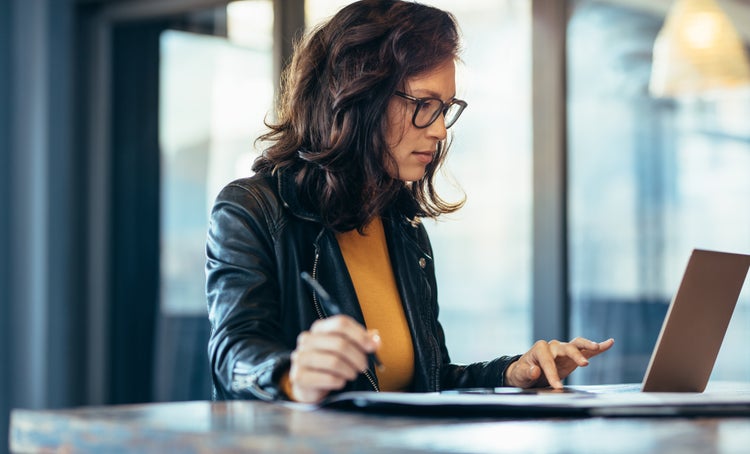 A woman wearing a leather jacket and glasses uses her laptop to learn how invoice processing works.