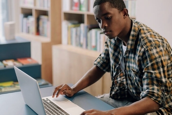 A college student sitting at a desk using their laptop in a library