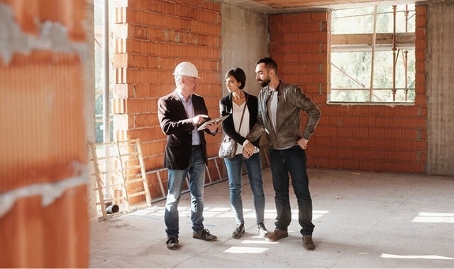An image of three people standing inside a room at a construction site and talking. One of the people is holding a tablet and pointing to it.