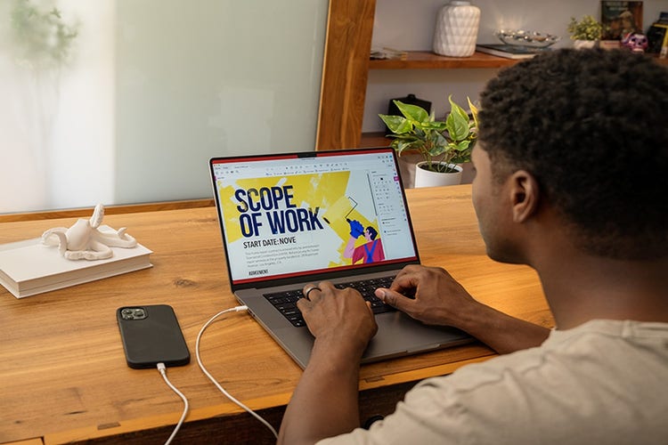 Person sitting at a woden desk with a cellphone and a laptop, flattening a PDF for a ‘scope of work’ presentation.