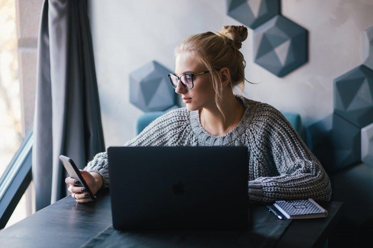A woman checks her phone while using a Mac to convert HEIC files to JPG.