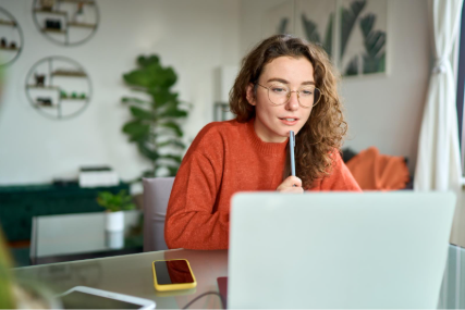A young woman looks at a laptop screen with a pen held to her chin in deep thought.