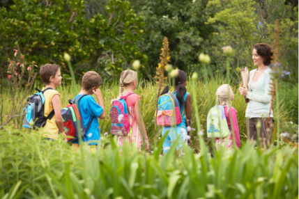 Teacher leading a group of elementary school children on a nature field trip, with kids wearing colorful backpacks and walking through a lush green outdoor setting.