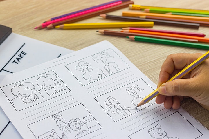 A hand holding a pencil adding drawings to a storyboard template with other colored pencils in the frame.