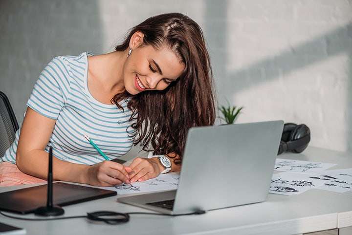 Young woman using a pencil at a desk on a series of drawings in front of a computer screen.