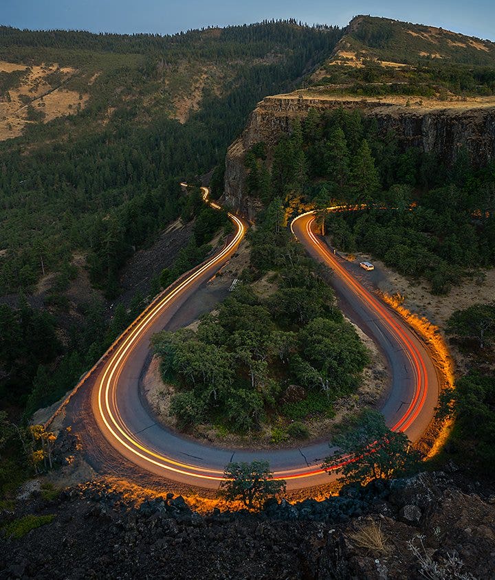 An aerial motion blur photo of cars travelling on a road that runs through a forest hillside
