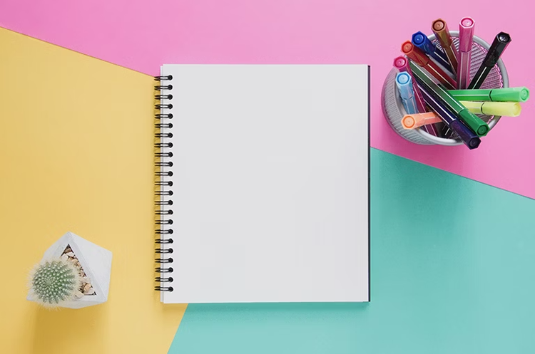 Blank spiral notebook on a pink, yellow and green background, with a pot of coloured pens and a small cactus ornament nearby