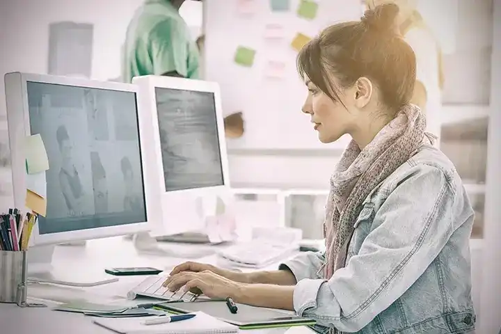 Woman working at a dual-monitor desk setup, with colleagues and sticky notes visible in the background