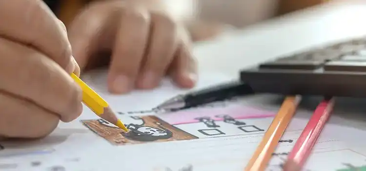 Close-up of hands colouring a character illustration with a yellow pencil, surrounded by coloured pencils and a keyboard on a desk